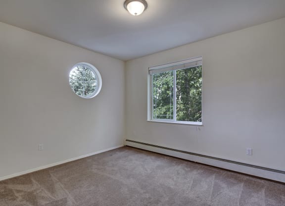 An empty room here at The Highlands Luxury Apartments with beige carpet, white walls, and two windows. A round window shows a tree outside, and a larger window has blinds, offering greenery views.