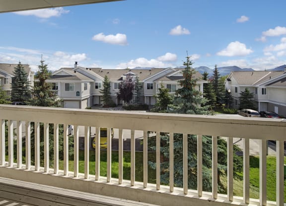 View from a balcony here at The Highlands Luxury Apartments overlooking a row of townhouses with a bright blue sky and scattered clouds. Tall pine trees and parked cars line the street below.