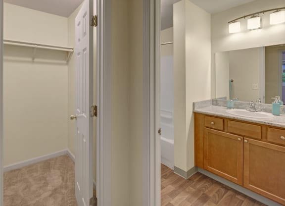 A modern bathroom here at Hillside Chalet Apartments features a spacious vanity with a marble countertop, wooden cabinets, and dual sinks. Adjacent is a carpeted closet space.