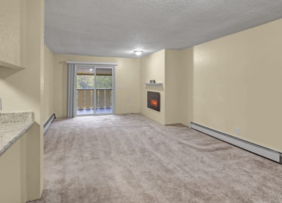 Empty living room here at Hillside Chalet Apartments with beige walls and carpet, featuring a corner electric fireplace. Sliding glass door leads to a balcony with green foliage outside.