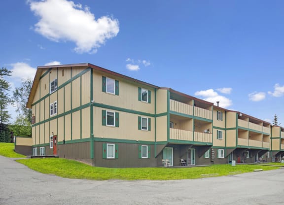 Three-story yellow and green Hillside Chalet apartment building with balconies, set against a bright blue sky. The foreground shows a grassy area and parking lot.