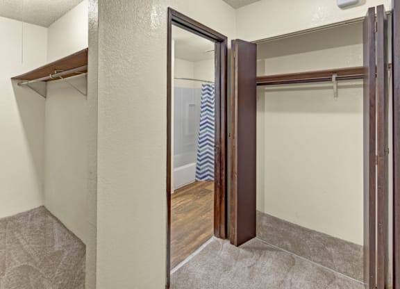 A small, empty walk-in closet here at Hillside Chalet Apartments with beige walls, open wooden shelves, and brown carpet. Leads to a bathroom with a chevron-patterned shower curtain.