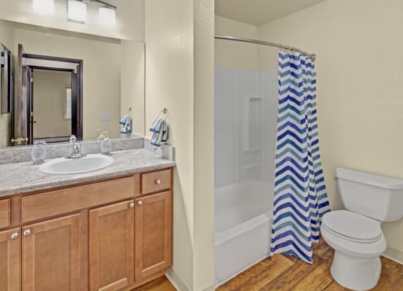 Bathroom here at Hillside Chalet Apartments with wooden cabinets, granite countertop, and mirror above a sink. A shower with a blue chevron curtain and a toilet beside it. Cozy ambiance.
