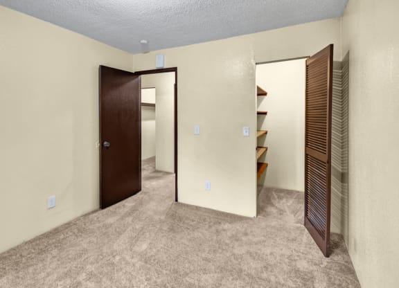 Empty beige bedroom here at Hillside Chalet Apartments with brown carpet, featuring an open door leading to another space with shelving. The atmosphere is simple and neutral.