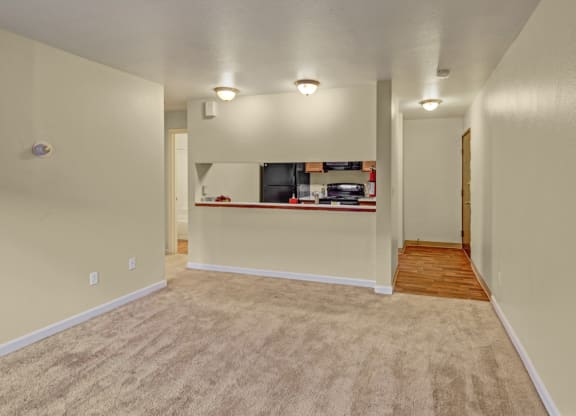 Quiet apartment interior here at Hillside Chalet Apartments with beige carpeted living area leading to a small kitchen. Warm lighting creates a cozy and inviting atmosphere.