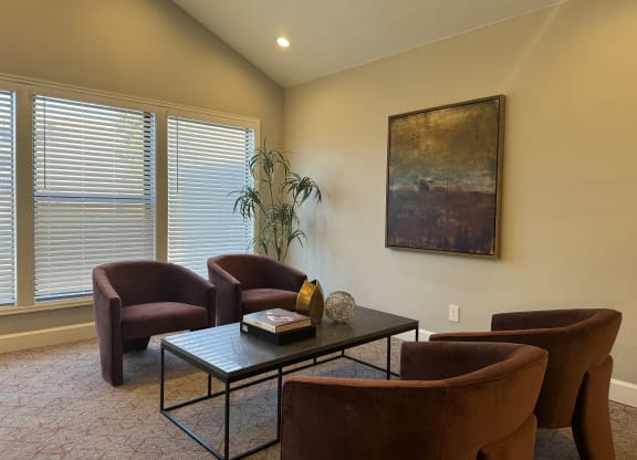 A cozy seating area here at Eagle Ridge Apartment Homes with four brown chairs around a black coffee table. A large abstract painting and a tall plant decorate the warm, well-lit room.