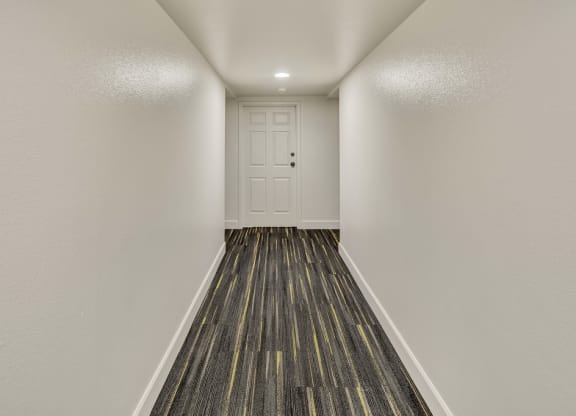 Narrow, brightly lit hallway at La Maisonnette Apartments with white walls and door at the end. The floor features dark patterned carpeting with subtle yellow stripes, creating a modern feel.