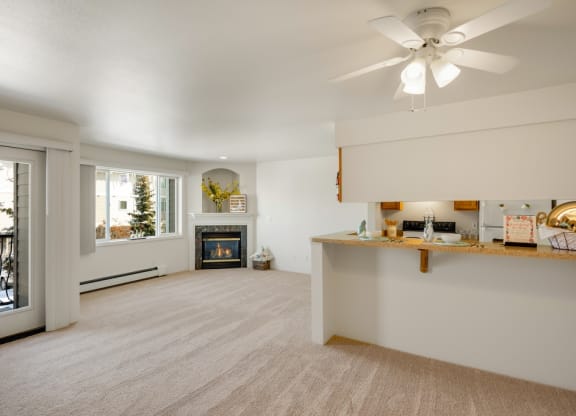 Bright living room here at La Maisonnette Apartments with beige carpet, a fireplace, large windows, and a kitchen bar. Soft, neutral tones create a cozy, welcoming atmosphere.