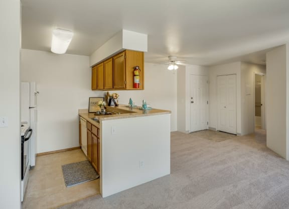 Cozy apartment interior here at La Maisonette Apartments featuring a small kitchen with wooden cabinets, beige countertops, and a dining bar. Adjacent is a carpeted living space, with closed white-paneled doors and visible lighting, creating a warm and inviting atmosphere.