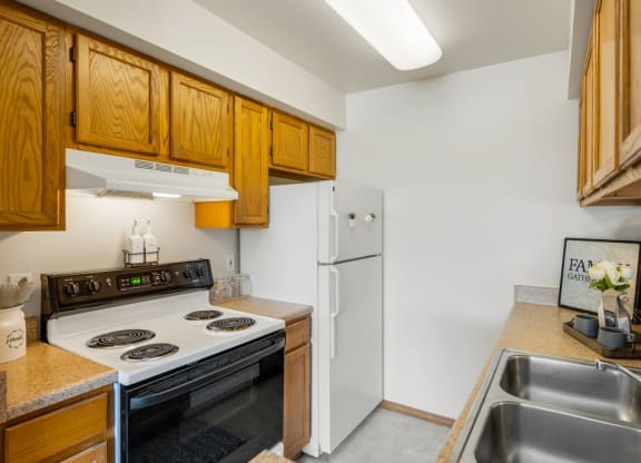 Compact kitchen here at La Maisonnette Apartments with wooden cabinets, a white stove, and fridge. Stainless steel sink in the foreground, decor includes a “Family” sign and flowers.