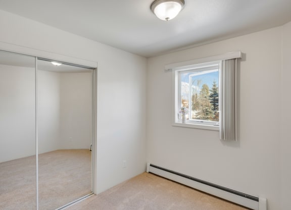 Small bedroom here at La Maisonnette Apartments with beige carpet and white walls, featuring a window with vertical blinds, a ceiling light, and a mirrored closet door reflecting light.