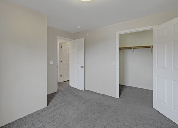 Empty bedroom here at La Maisonnette Apartments with gray carpet and light beige walls. Two open doors reveal a closet and a hallway. The space feels clean and minimalistic.