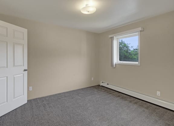 Bedroom here at La Maisonnette Apartments with beige walls, a gray carpet, and a white door ajar. A window with blinds lets natural light in, creating a calm, neutral ambiance.
