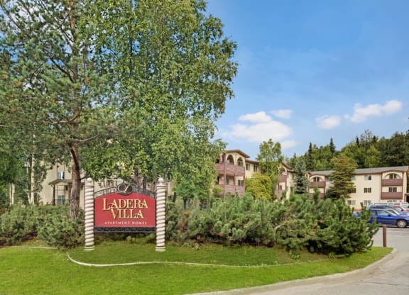 A serene apartment complex with a sign reading "Ladera Villa" amidst lush greenery and tall trees. The buildings are cream with arched windows. The sky is clear.