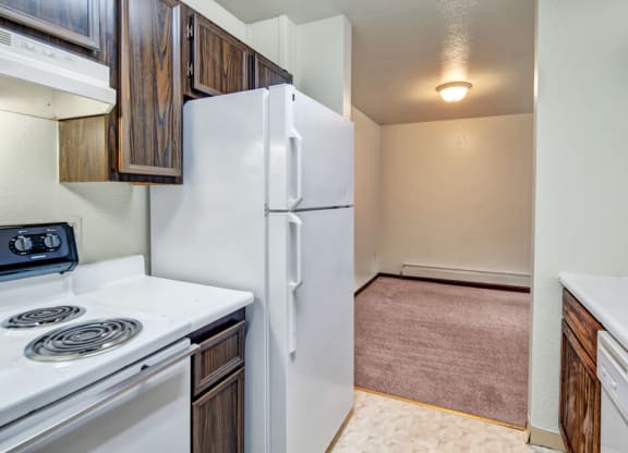 Compact kitchen here at Ladera Villas Apartments with wooden cabinets, white stove and fridge. Leads to a carpeted dining area, softly lit by a round ceiling light. Cozy and simple.