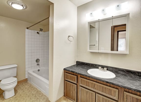 A tidy bathroom here at Ladera Villas Apartments with a bathtub-shower combo, white tiles, and a white toilet. A wooden vanity with a marble countertop sits beneath a lit mirror.