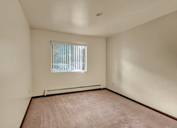 Bedroom here at Ladera Villas Apartments with beige carpet and cream walls, lit by sunlight through vertical blinds on a window. The tone is warm and inviting.