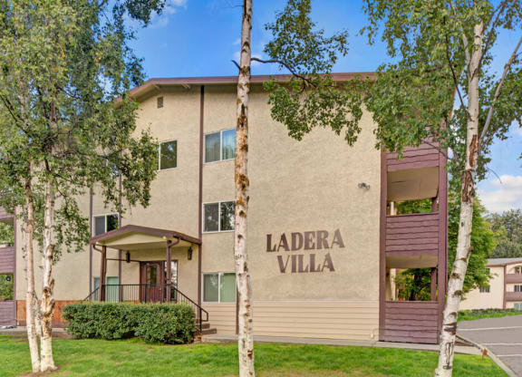 Three-story beige Ladera Villa apartment building labeled "Ladera Villa" with brown accents. Surrounded by lush green trees and grass under a clear blue sky.