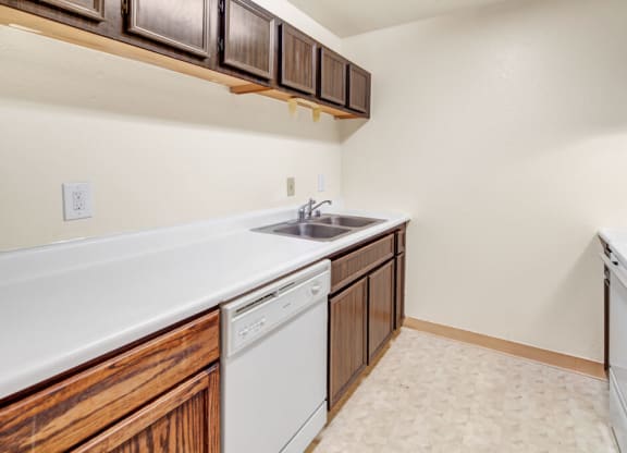 A narrow kitchen here at Ladera Villas Apartments with dark brown cabinets, white countertops, and a stainless steel sink. A dishwasher is below, and the room has a neutral, minimalist tone.