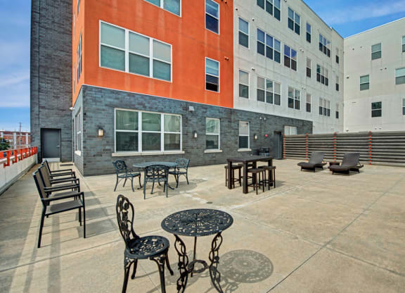 Patio at the Latitude Apartments with metal tables and chairs, loungers, and a barbecue grill. Surrounding buildings feature orange and grey facades under a clear sky.