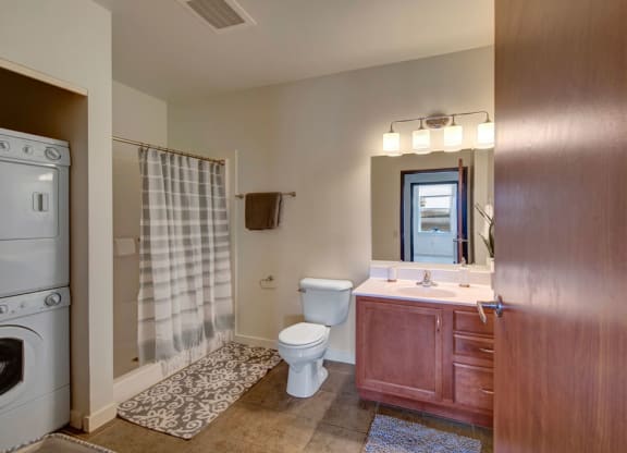 Modern bathroom at the Latitude Apartments with light wood cabinetry, a large mirror, and warm lighting. A striped shower curtain is adjacent to a stacked washer and dryer.