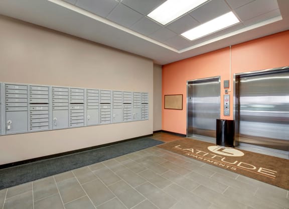 The Latitude Apartment lobby with gray mailboxes on a beige wall, two closed elevators on an orange wall, and a brown mat reading "Latitude Apartments" on tiled flooring.