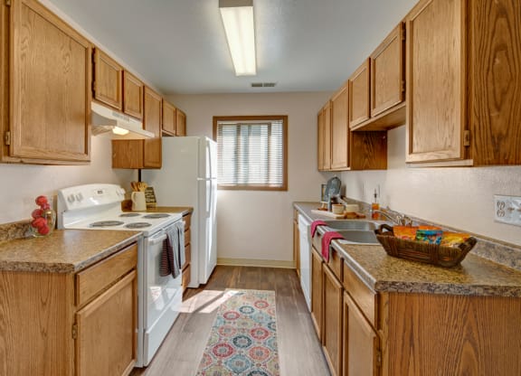 Narrow kitchen here at Legacy West Apartments with wood cabinets, white appliances, and granite countertops. A rug adds warmth, while natural light streams through a window. Cozy ambiance.
