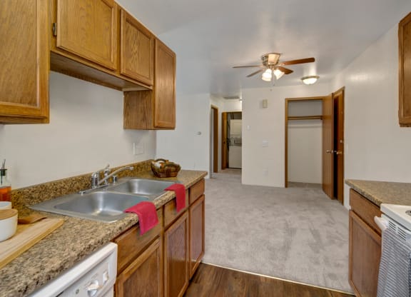 Kitchen here at Legacy West Apartments with wood cabinets and granite countertops, double sink with red towel, leading to carpeted hall. Ceiling fan and open closet visible. Cozy ambiance.