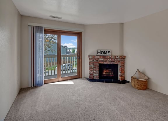 Empty living room here at Legacy West Apartments with a cozy brick fireplace, sign reading "Home," and a wicker basket on plush carpet. Sunlit balcony doors reveal trees outside.