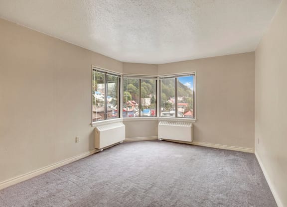 Living room here at Mendenhall Tower Apartments with beige walls and gray carpet, featuring a bay window. The window offers a view of colorful houses and lush green hills under a blue sky.