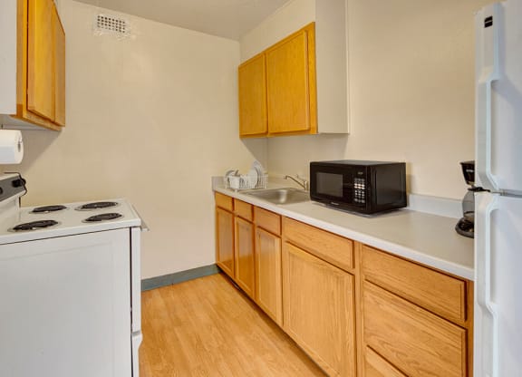 Compact kitchen here at Mendenhall Tower Apartments with light wood cabinets, white appliances, and a microwave. Includes an electric stovetop, sink with dish rack, and a coffee maker.