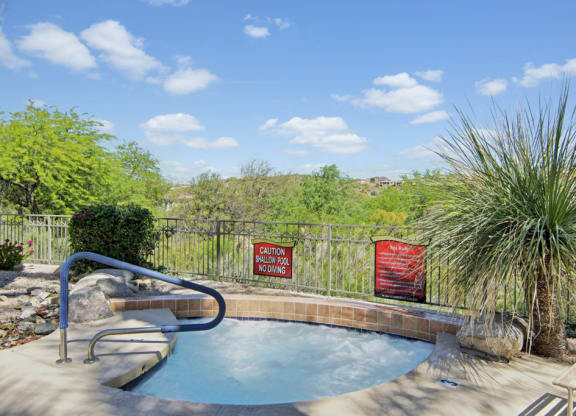 Outdoor hot tub here at Mirador Apartments surrounded by greenery and a metal fence under a blue sky with fluffy clouds. Warning signs are visible near the tub edge.