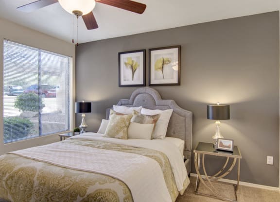 A cozy bedroom here at Mirador Apartments with a plush bed adorned with patterned pillows and a beige comforter. Two framed botanical prints hang on a gray wall. Soft lighting from two bedside lamps and a window add warmth. A ceiling fan is visible above.