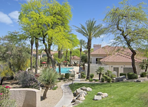 Sunny courtyard here at Mirador Apartments with vibrant greenery, palm trees, and well-maintained landscape. Central pathway leads to a swimming pool near a beige house with a red-tiled roof.