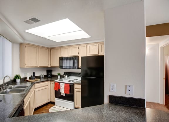 Bright kitchen Mirador Apartments with light wood cabinets, black appliances, and dark granite countertops. Decor includes potted plants and red towels. Cozy and modern feel.