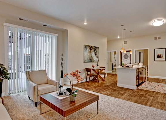 A welcoming open-concept living area here at 4800 Excelsior featuring a comfortable seating arrangement with a lounge chair and coffee table set on soft carpeting beside large vertical windows. The space flows into a dining nook and modern kitchen with an island, pendant lighting, and warm wood-style flooring, creating a bright and inviting home layout.