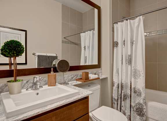 A modern bathroom here at 4800 Excelsior featuring a stone countertop vanity with an undermount sink, a framed wall mirror, and neatly arranged accessories. Neutral tile surrounds the tub and shower with a patterned shower curtain, while warm cabinetry, soft lighting, and clean finishes create a polished, spa-inspired space.