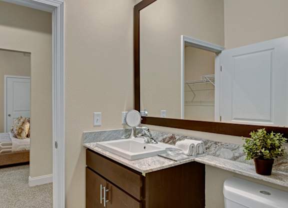 A stylish bathroom here at 4800 Excelsior featuring a stone countertop vanity with an undermount sink, warm wood cabinetry, and a large framed mirror. Open shelving holds neatly folded towels, while neutral walls and clean finishes create a bright, polished space with a connected view toward the bedroom and closet area.