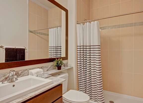 A clean bathroom here at 4800 Excelsior featuring a stone countertop vanity with an undermount sink, warm wood cabinetry, and a large framed mirror. A tiled tub and shower with a patterned curtain sits beside the vanity, while neutral tile walls, soft lighting, and neatly placed towels create a calm, well-organized space.