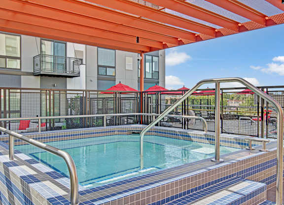 A modern outdoor spa here at 4800 Excelsior featuring a tiled hot tub with built-in steps and stainless handrails beneath a bold orange pergola. The fenced courtyard includes contemporary apartment façades, red patio umbrellas, and seating areas, creating a vibrant, inviting space for relaxation and socializing.
