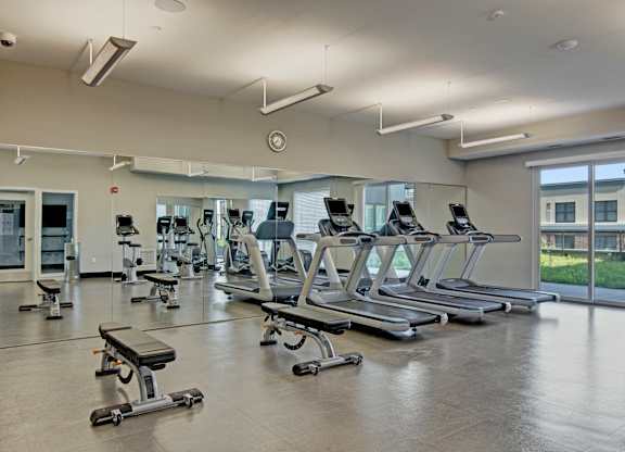 A modern fitness center here at 4800 Excelsior featuring multiple treadmills, weight benches, and cardio equipment arranged across a spacious open floor. Large wall mirrors and overhead lighting enhance the bright atmosphere, while tall windows bring in natural light and views of the surrounding community.