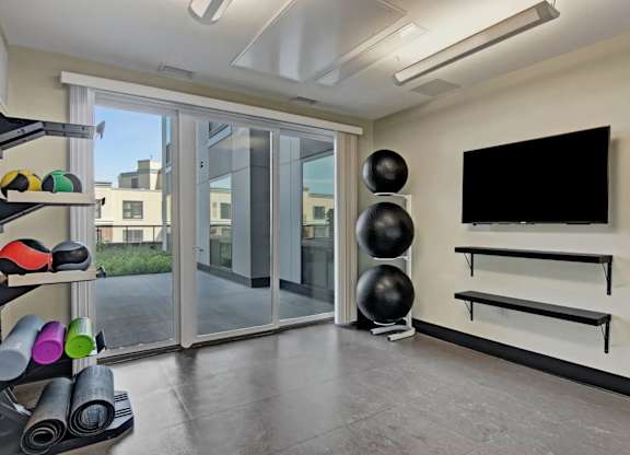 A dedicated fitness studio here at 4800 Excelsior showcasing an open workout space with stability balls, yoga mats, and fitness accessories neatly stored along the wall. Large sliding glass doors lead to an outdoor terrace and bring in natural light, while mounted shelving and a wall-mounted screen support guided workouts and training sessions.