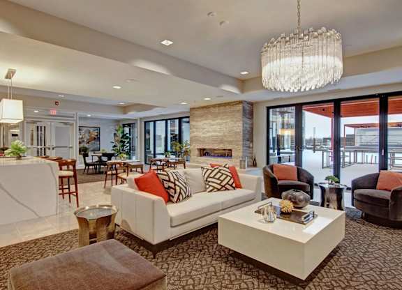 A stylish resident lounge here at 4800 Excelsior featuring a modern seating area with a white sofa, accent chairs, and decorative tables arranged around a contemporary fireplace. The space includes a large chandelier, a sleek bar with counter seating, and floor-to-ceiling windows that open to an outdoor terrace, creating an inviting area for relaxing or gathering.