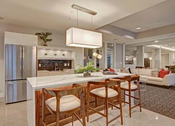 A modern community kitchen here at 4800 Excelsior featuring a large island with a smooth countertop, wood-accented cabinetry, and bar seating with upholstered stools. Stainless steel appliances, open shelving with greenery, and soft pendant lighting complement the adjacent lounge seating area, creating a welcoming space for cooking, dining, and socializing.