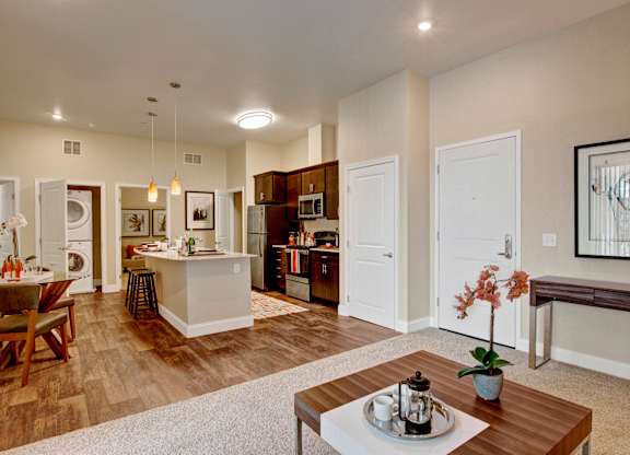 A bright open-concept living space here at 4800 Excelsior showcasing a carpeted seating area that flows into a modern kitchen with dark cabinetry, stainless appliances, and a central island with pendant lighting. Wood-style flooring defines the kitchen and dining areas, while neutral walls, framed artwork, and thoughtful decor create a warm, cohesive apartment layout.