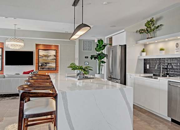 A contemporary community kitchen here at 4800 Excelsior featuring a spacious waterfall island with bar seating, sleek white cabinetry, and a modern tile backsplash. Stainless steel appliances, open shelving with greenery, and stylish pendant lighting complement the adjacent lounge area, creating a bright and inviting space for gatherings and everyday use.