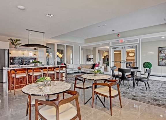 A stylish community lounge here at 4800 Excelsior featuring round café tables with upholstered chairs, a spacious kitchen island with bar seating, and a comfortable seating area with sofas and accent chairs. Large windows, modern lighting, and sleek finishes create an inviting space for socializing, working, or relaxing.