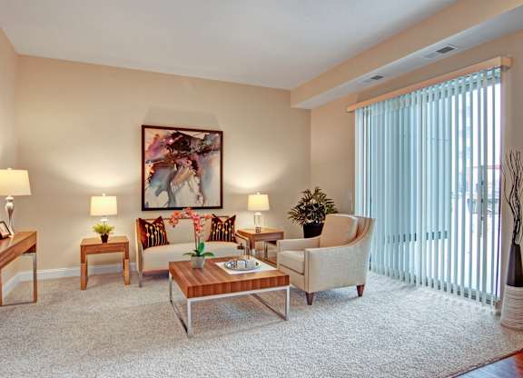A warm living room here at 4800 Excelsior featuring plush carpeting, a comfortable sofa with accent pillows, a matching armchair, and a wooden coffee table centered on the space. Coordinated side tables with lamps, abstract wall art, and a sliding glass door with vertical blinds create a bright, inviting setting with natural light and thoughtful details.