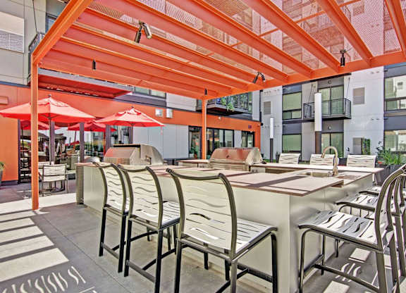A modern outdoor grilling area here at 4800 Excelsior featuring a spacious built-in grill station with bar-style seating beneath a bold orange pergola. Sleek countertops, high-back chairs, and nearby patio tables with umbrellas create an inviting setting for gatherings, surrounded by contemporary apartment architecture and open-air courtyard views.