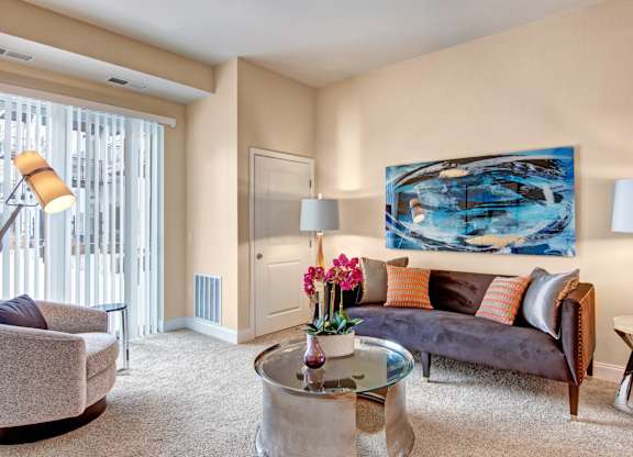 A cozy living room here at 4800 Excelsior featuring plush carpeting, a contemporary sofa with accent pillows, and a round glass coffee table centered in the space. Soft lighting, abstract wall art, and a large window with vertical blinds create a warm, comfortable atmosphere with plenty of natural light.